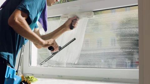 Person cleaning a large window with a squeegee, spray bottle, and towel on their shoulder.