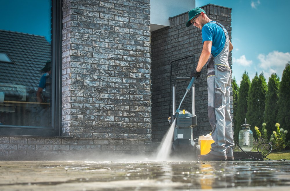 Man in a teal cap and blue shirt uses a pressure washer to clean a brick exterior wall of a house; water spray visible outside. The scene is sunny and outdoors.