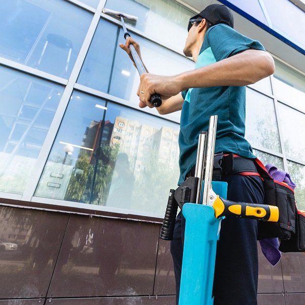 A maintenance worker in a teal shirt and cap cleans the exterior glass of a modern building, wearing a tool belt and carrying cleaning tools.