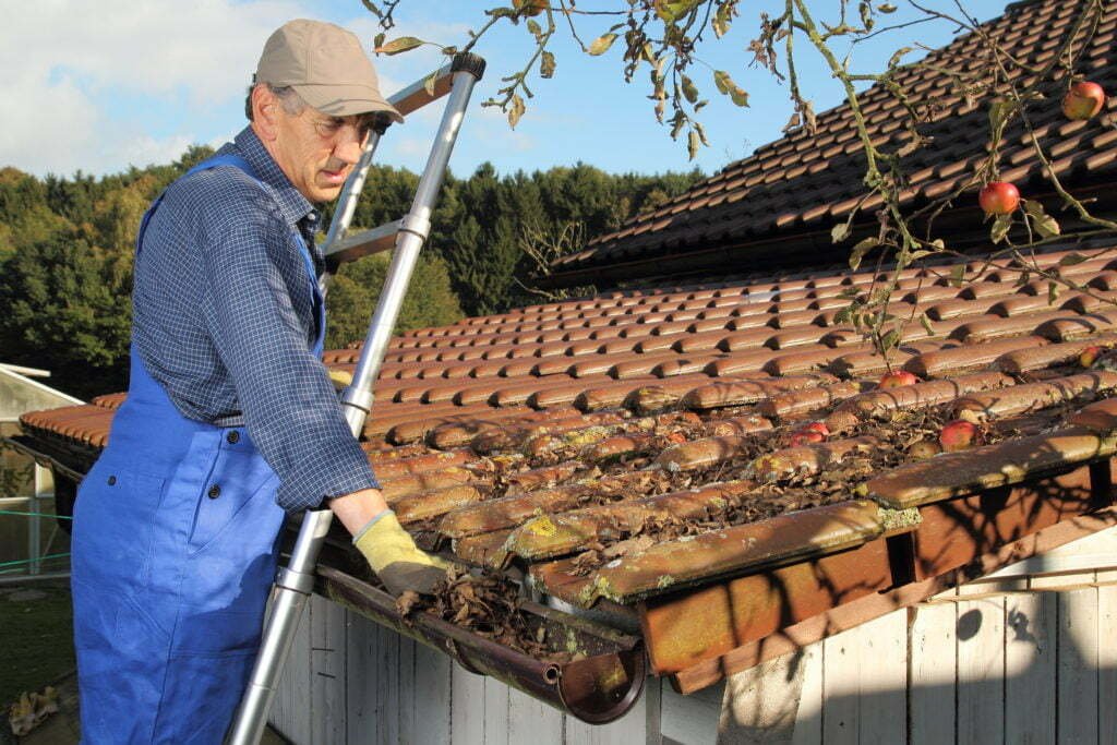 Older man in blue overalls cleaning leaves from terracotta roof tiles with a telescoping tool on a sunny day.
