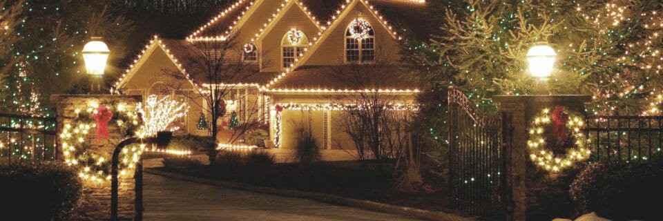 Two-story house at night wrapped in warm Christmas lights along roof, porch, and gate, with wreaths and lit trees nearby.