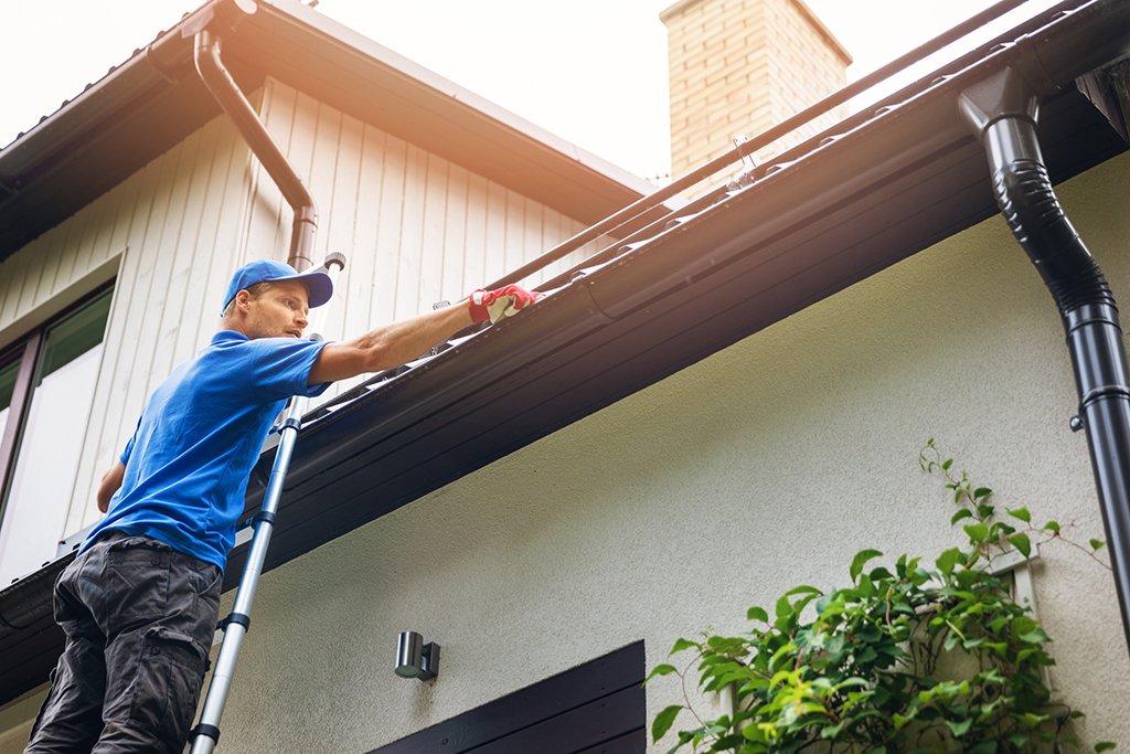 Man in a blue shirt and cap stands on a ladder, repairing or cleaning the house gutter along the roofline.