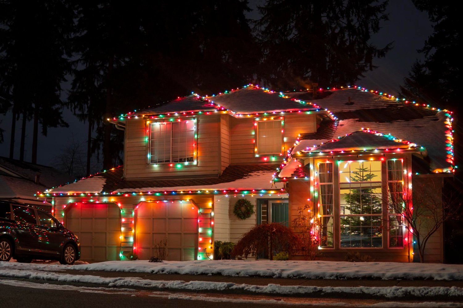 Two-story house decorated with multicolored Christmas lights along the roofline and windows at night; snow on the ground.