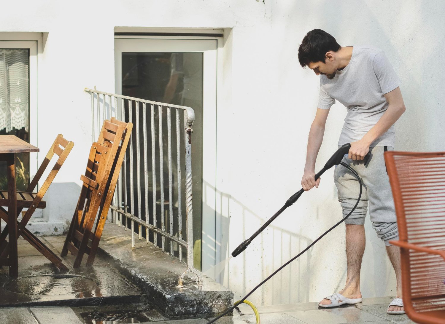 Man in a gray t-shirt and joggers pressure-washing a white building's balcony outdoors, with wooden chairs nearby.
