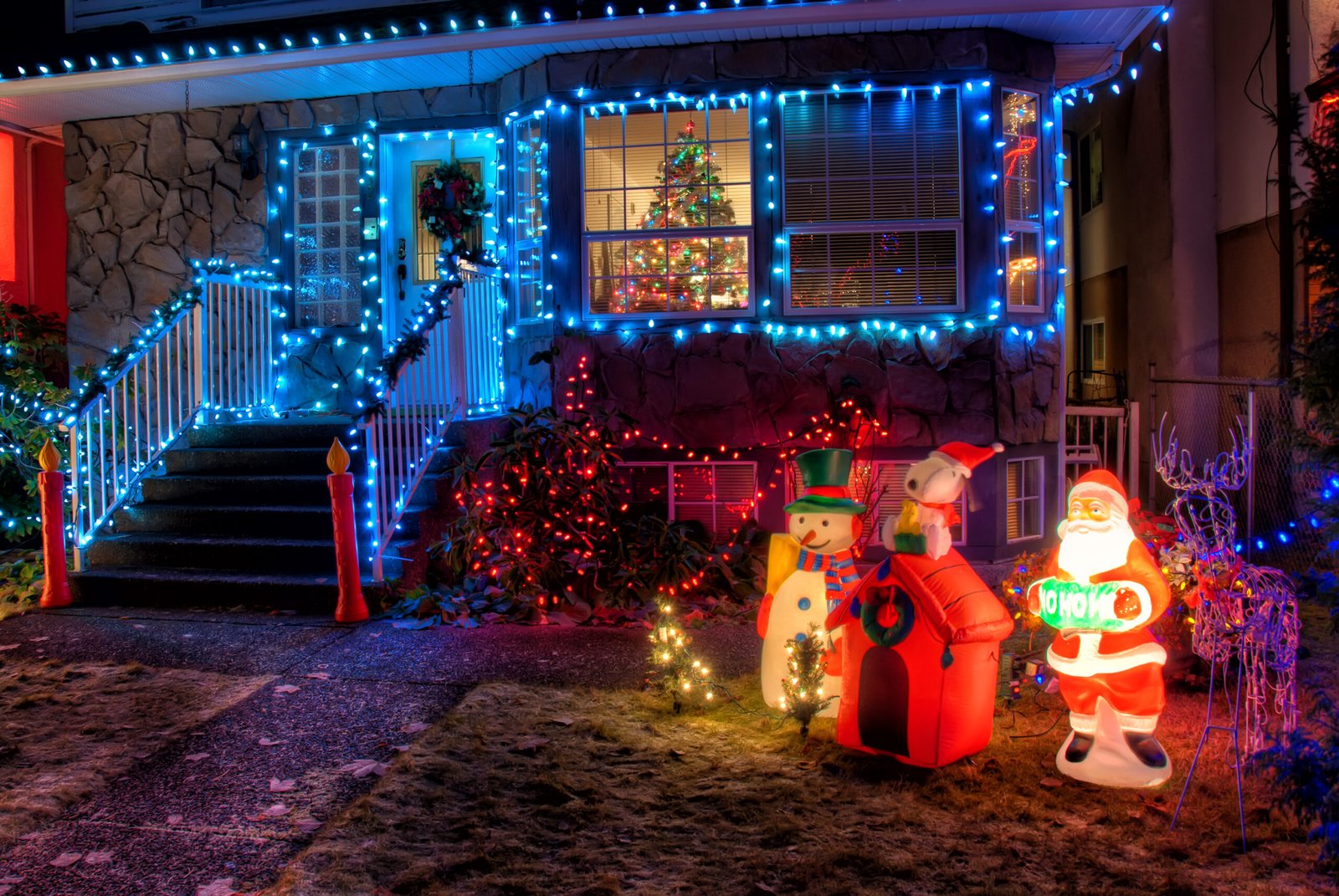 House outlined with blue Christmas lights on stairs and windows; inflatable Santa and snowman in the yard at night.