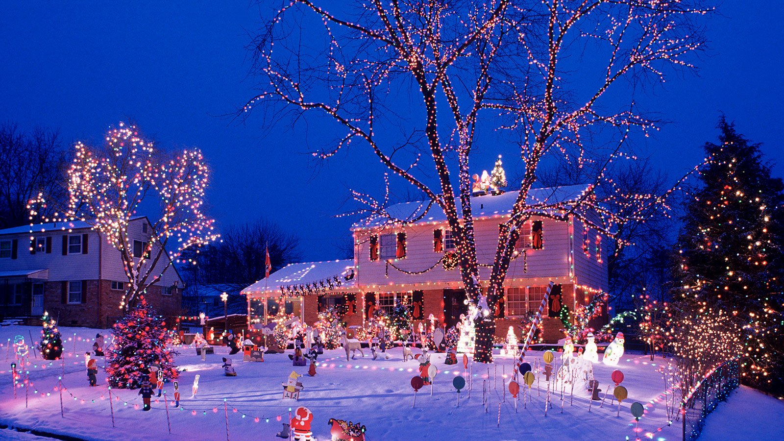 Snowy suburban yard at night adorned with abundant Christmas lights on the house, trees, and yard decorations.