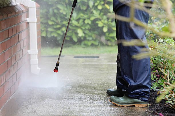 Person using a pressure washer to clean a concrete walkway beside a brick wall, wearing green boots and dark rain pants.