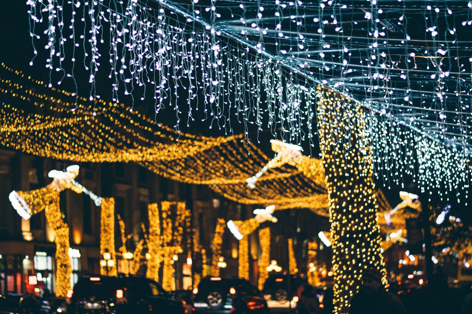 Night street decorated with warm yellow string lights on trees and blue-white hanging lights overhead, creating a festive glow at night