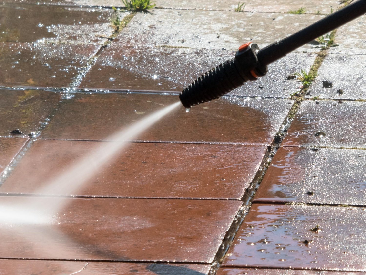 A spray from a pressure washer nozzle cleaning brown patio tiles.