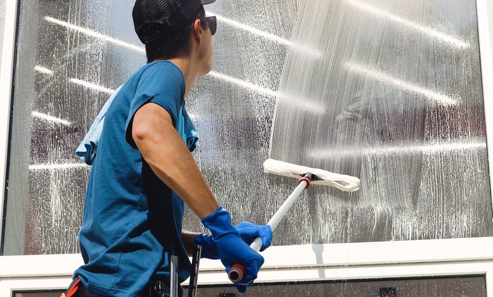 Person wearing a blue shirt and gloves cleans a large window with a squeegee outside.