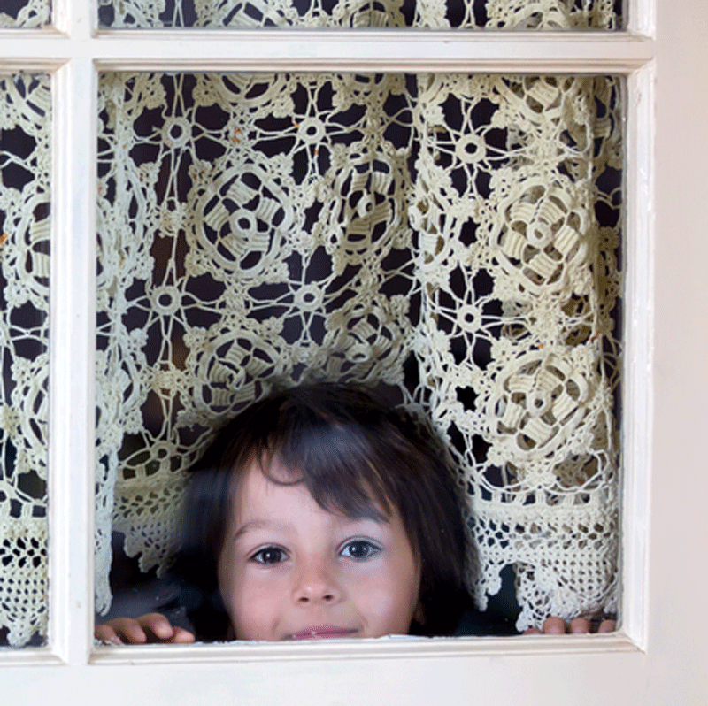 Child peeking through a window framed by intricate white lace curtains, hands resting on the sill.
