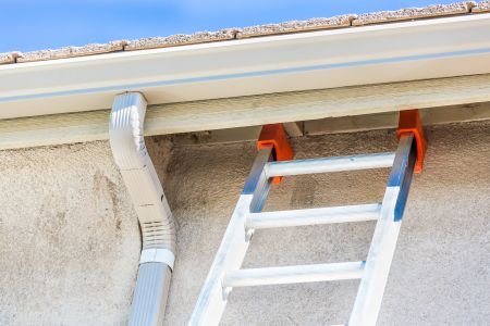 Aluminum ladder leaning against a stucco wall under the eave, near a white gutter, set up for roof work.