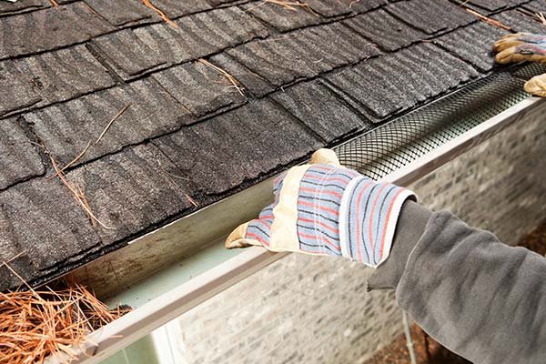 Hands in striped gloves fitting a metal gutter screen along the edge of a shingled roof.