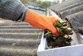 Pair of gloved hands cleaning leaves out of a roof gutter or drainage channel on a corrugated roof.