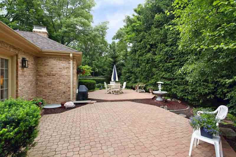 Backyard patio with brick house on the left, circular table with umbrella, and surrounding green hedges and trees.
