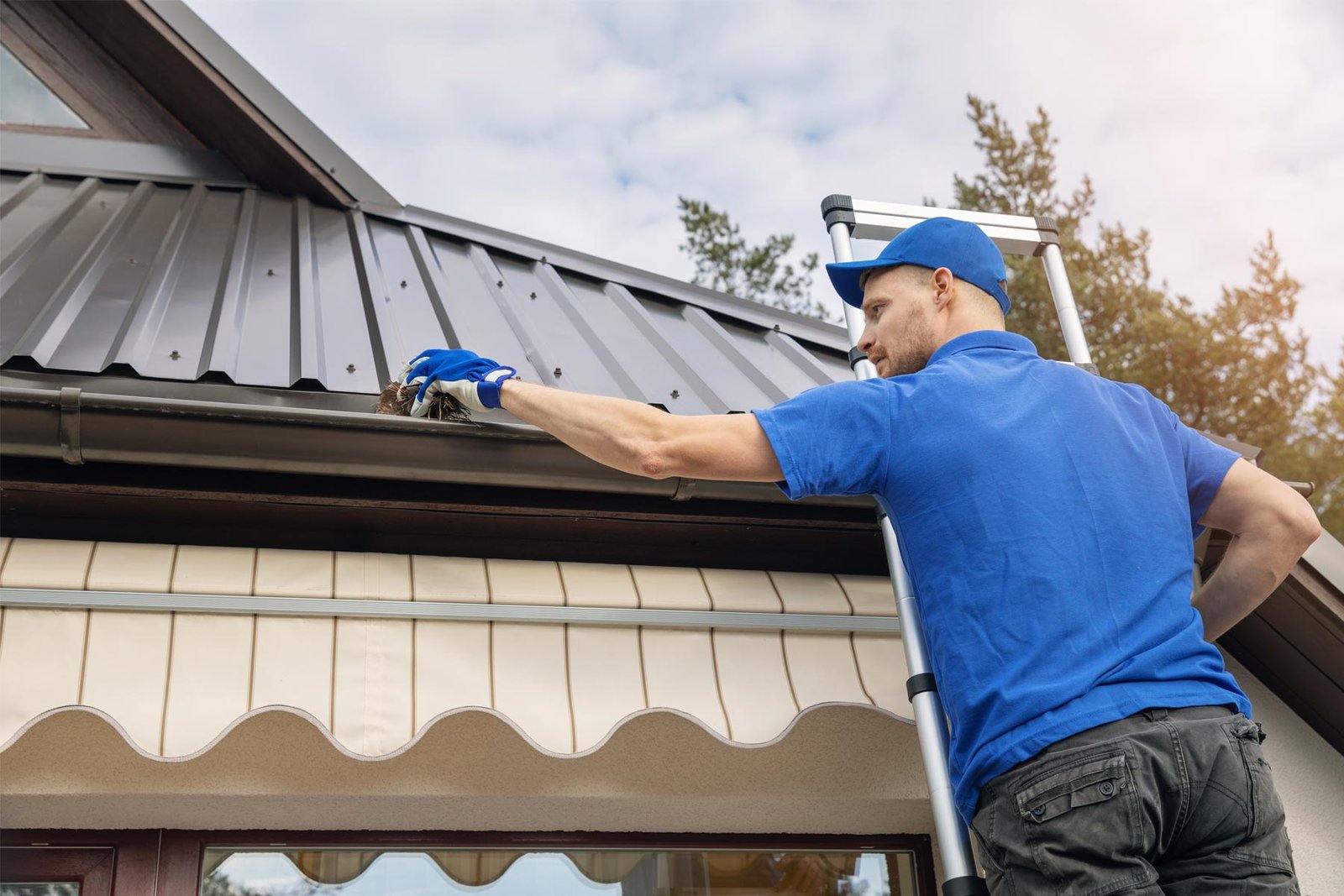 Man in a blue uniform on a ladder cleaning debris from a metal roof gutter.