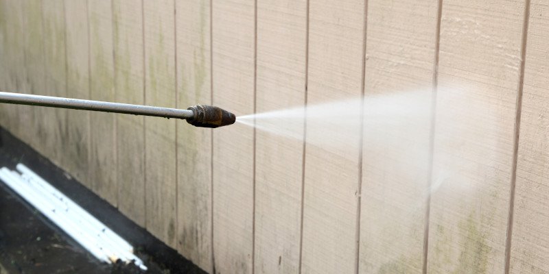 Pressure washer nozzle spraying a jet of water along a beige wooden wall for cleaning.