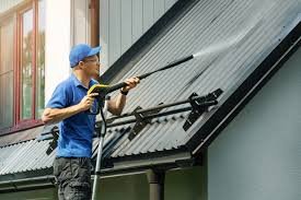 Man in a blue shirt and cap using a pressure washer to clean a sloped metal roof.