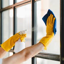 Person wearing yellow rubber gloves sprays cleaner and wipes a window with a blue microfiber cloth.