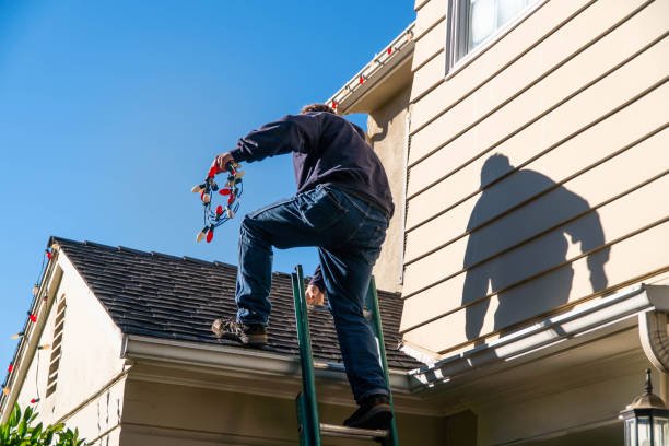 Person on a ladder decorating the beige house with colorful Christmas lights under a clear blue sky.