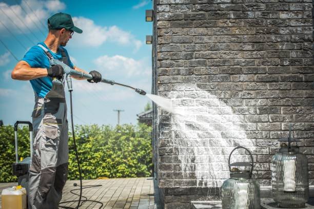 Man in work clothes pressure washing a brick wall outdoors, water spray arcing from the nozzle.
