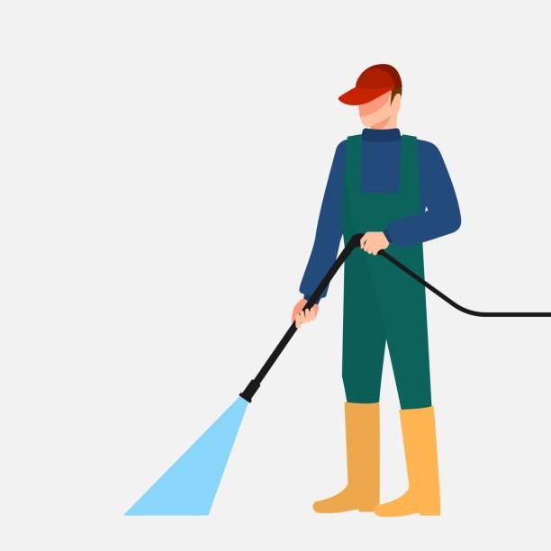 Worker in green overalls and yellow boots operating a pressure washer, spraying water onto a surface.