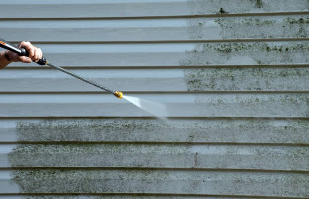 Person holding a pressure washer wand, spraying water to clean gray siding with moss and dirt.