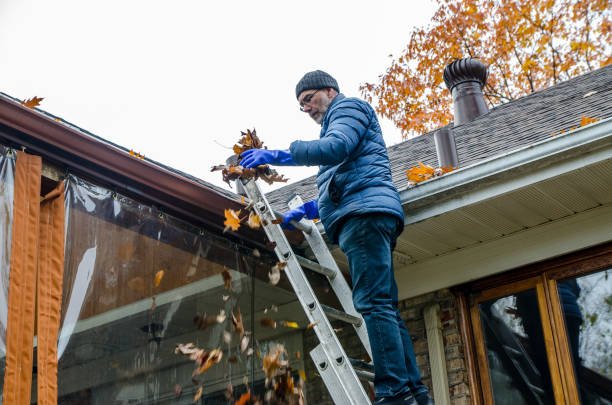 Man on a ladder cleaning fallen leaves out of a gutter on a fall day, tossing leaves into the air beside a house.