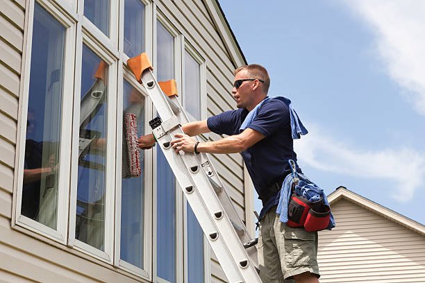 Man on a ladder washing the exterior window panes of a beige house, wearing a tool belt and sunglasses.