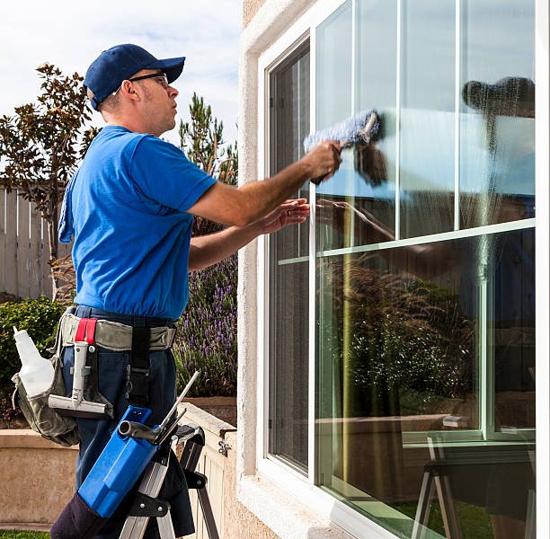 Man in a blue shirt cleans an exterior window with a squeegee, wearing a tool belt outdoors on a sunny day.