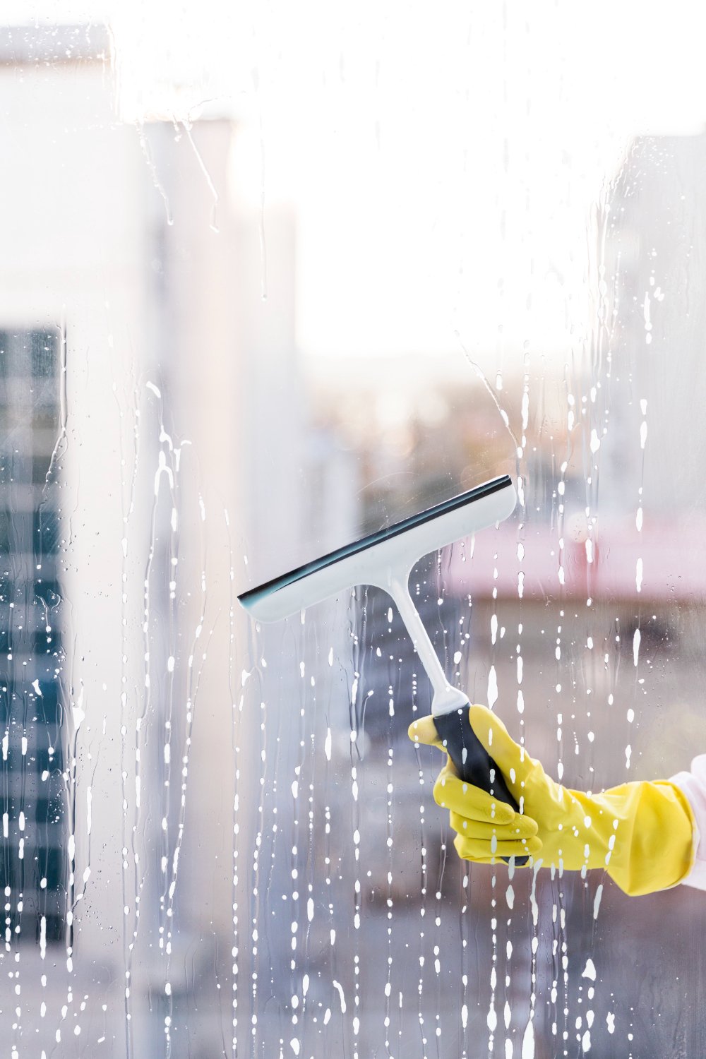 Person wearing yellow gloves uses a squeegee to wash a glass window, with city buildings blurred in the background (outdoors).Note: concise informative alt text for cleaning window image.