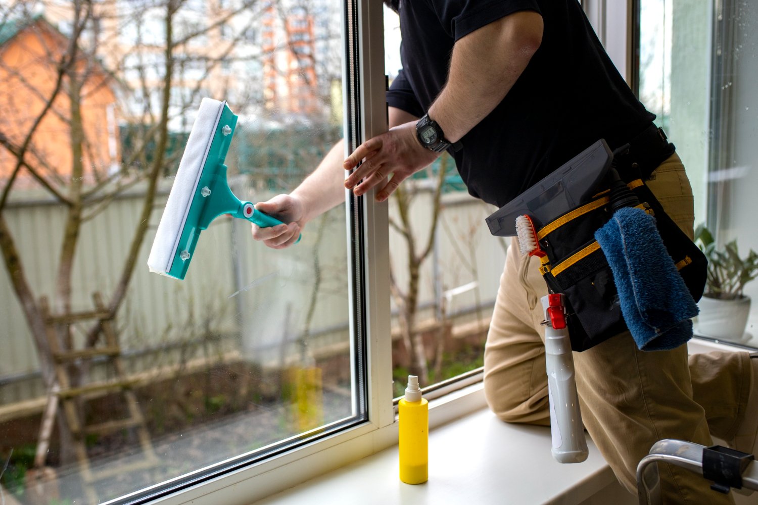 A window cleaner wearing a tool belt wipes a large glass window with a teal squeegee, sunlight outside and cleaning supplies nearby on the sill.