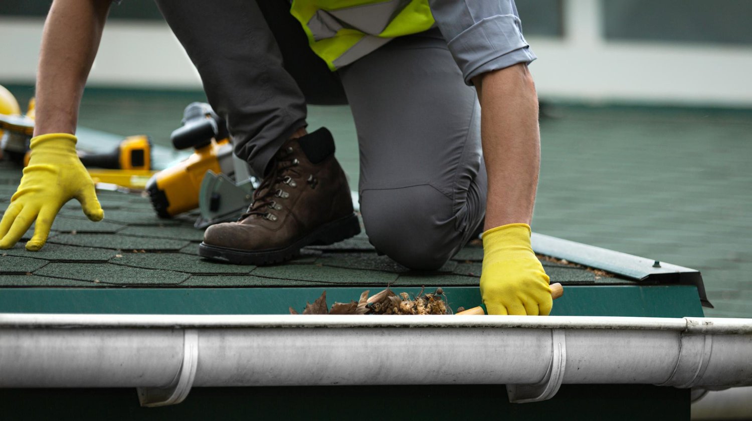 Worker in a high-visibility vest and yellow gloves kneels on a roof, clearing leaves with a nearby power tool and shingles visible ahead.