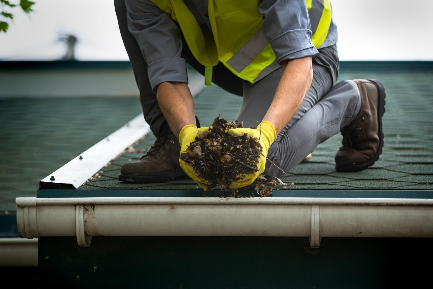 Maintenance worker in a safety vest and gloves kneels on a roof, gathering leaves and debris from the gutter edge side.
