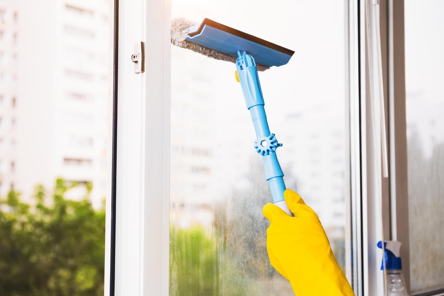 Person wearing a yellow glove uses a blue window squeegee to wipe a glass pane near a window frame.