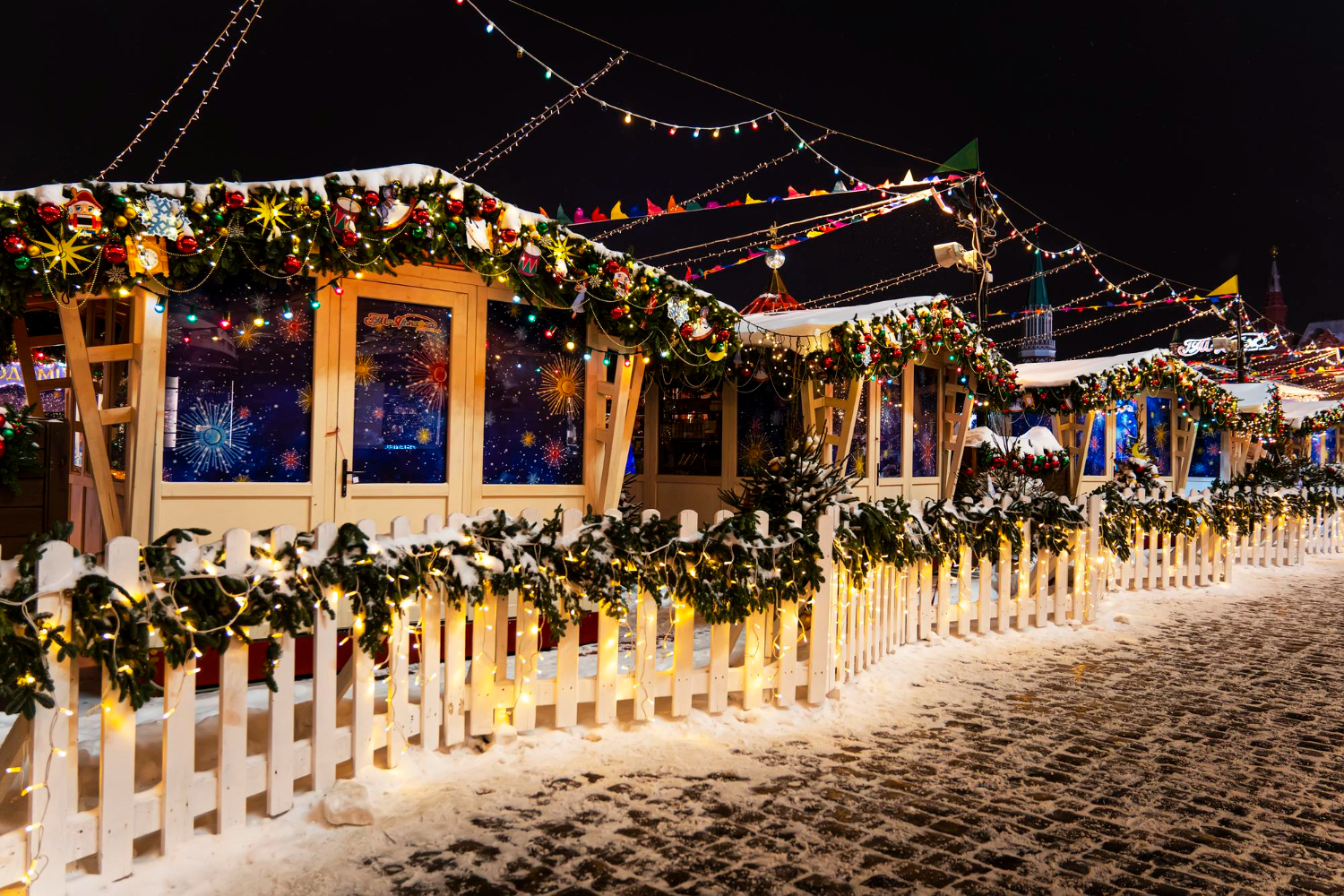 Row of festive wooden market stalls along a snow-covered street at night, decorated with garlands and colorful lights.