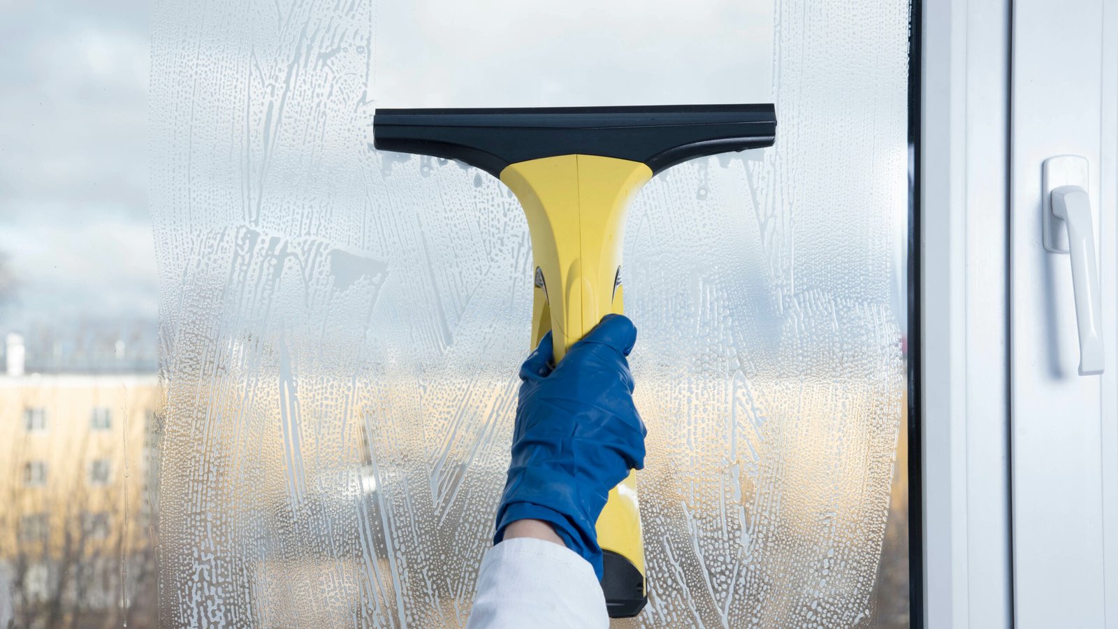 Hands in blue gloves holding a yellow squeegee to clean a soapy glass window pane.