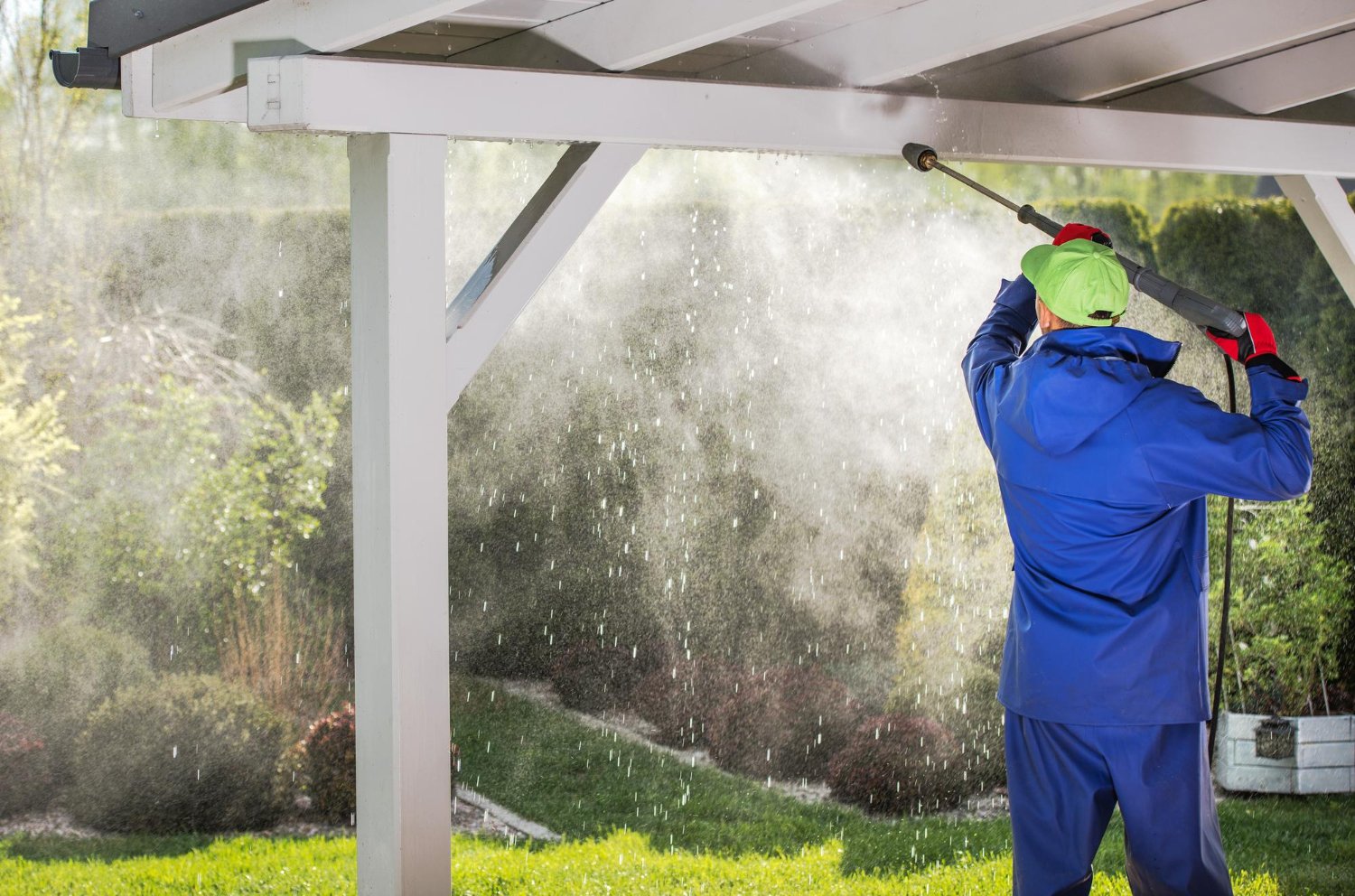 Maintenance worker in a blue rain suit and lime-green cap using a pressure washer under a white pergola, spraying water into a garden.