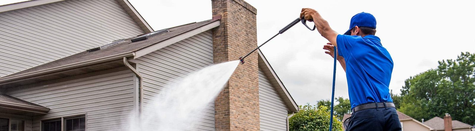 A man in a blue shirt and cap power-washes the side of a house with a high-pressure spray.