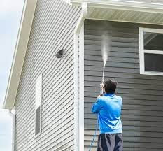Man pressure-washing the siding of a gray house, spraying water upward along the exterior