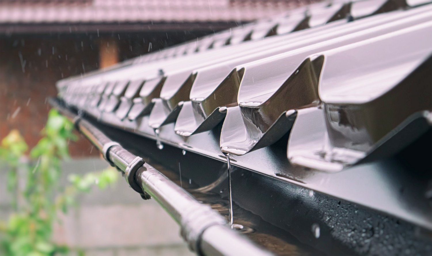 Close-up of a glossy metal roof with jagged edge tiles and falling rain, a downspout running along the edge below.