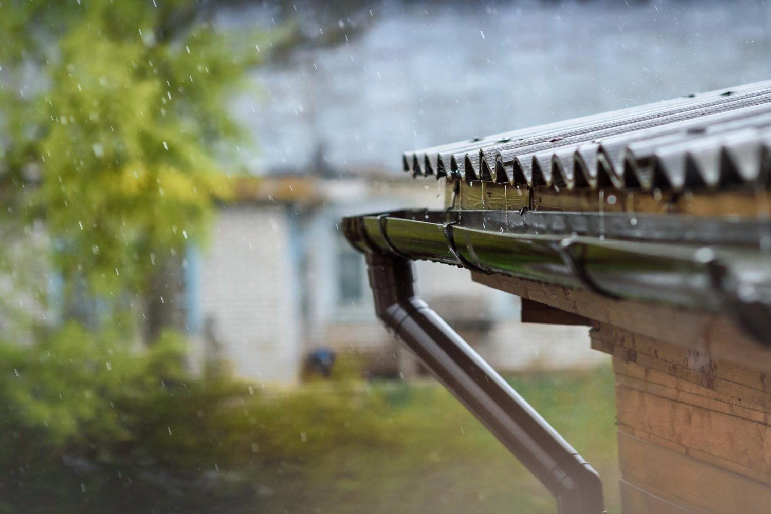 Rain falls on a metal gutter and downspout attached to a wooden house, with a blurred green yard in the background.