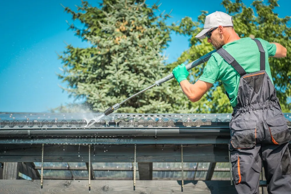 Man in green shirt and overalls power-washes a metal roof, spraying water across tiles outdoors on a sunny day with trees in the background