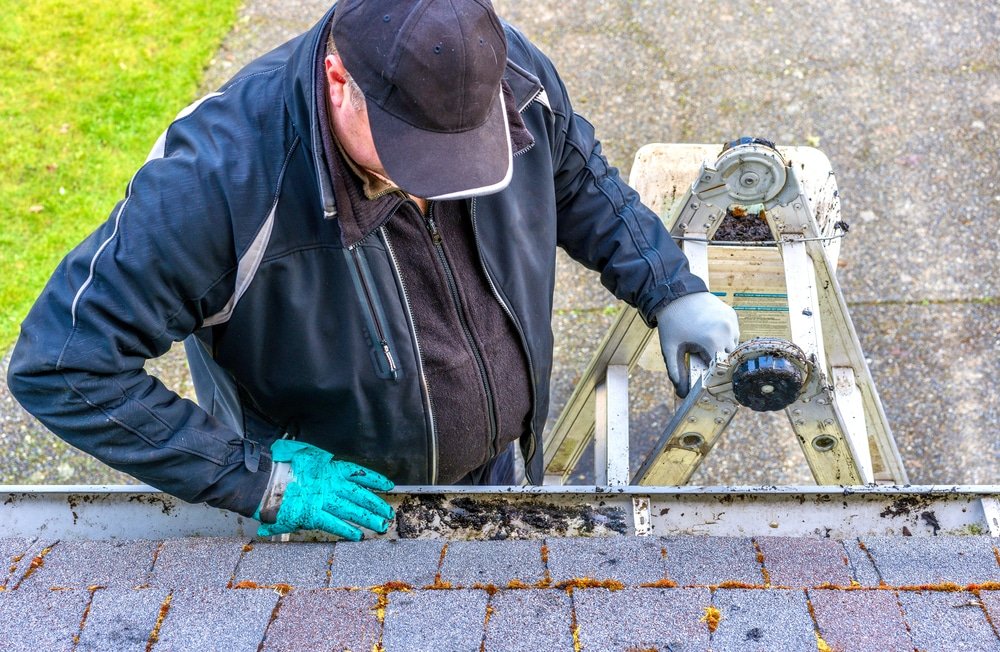 Man on a ladder cleaning debris from a gutter along a shingled roof in a residential setting.