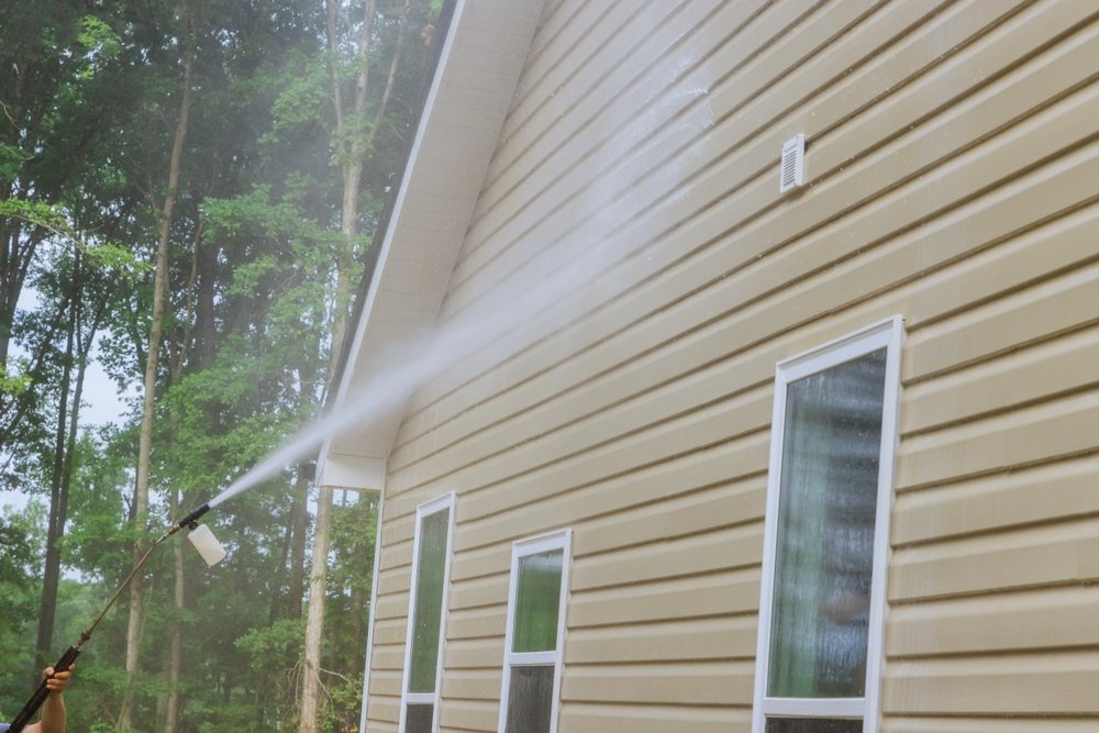 Person using a pressure washer to clean beige siding on a house, spray arc directed at the wall near two windows, trees in the background