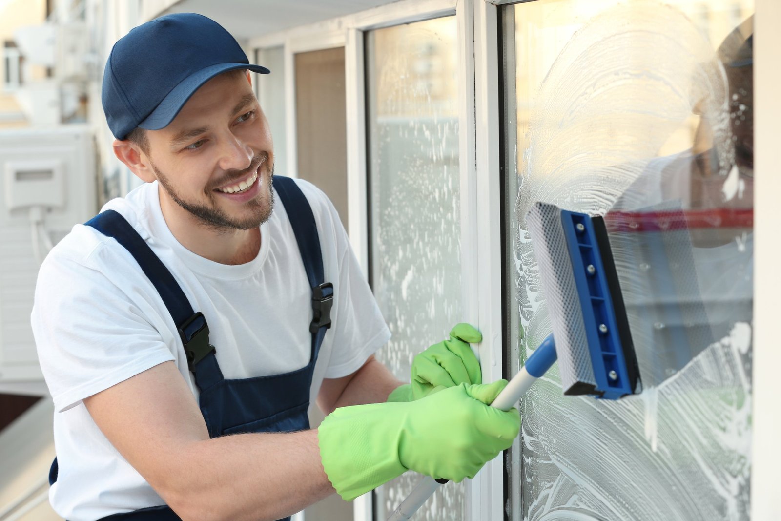 Smiling window cleaner in a white T-shirt and navy overalls wiping a glass door with a squeegee, wearing bright green gloves.