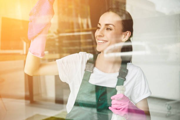 Smiling woman cleaning a window with a pink glove and cloth, wearing green overalls indoors.