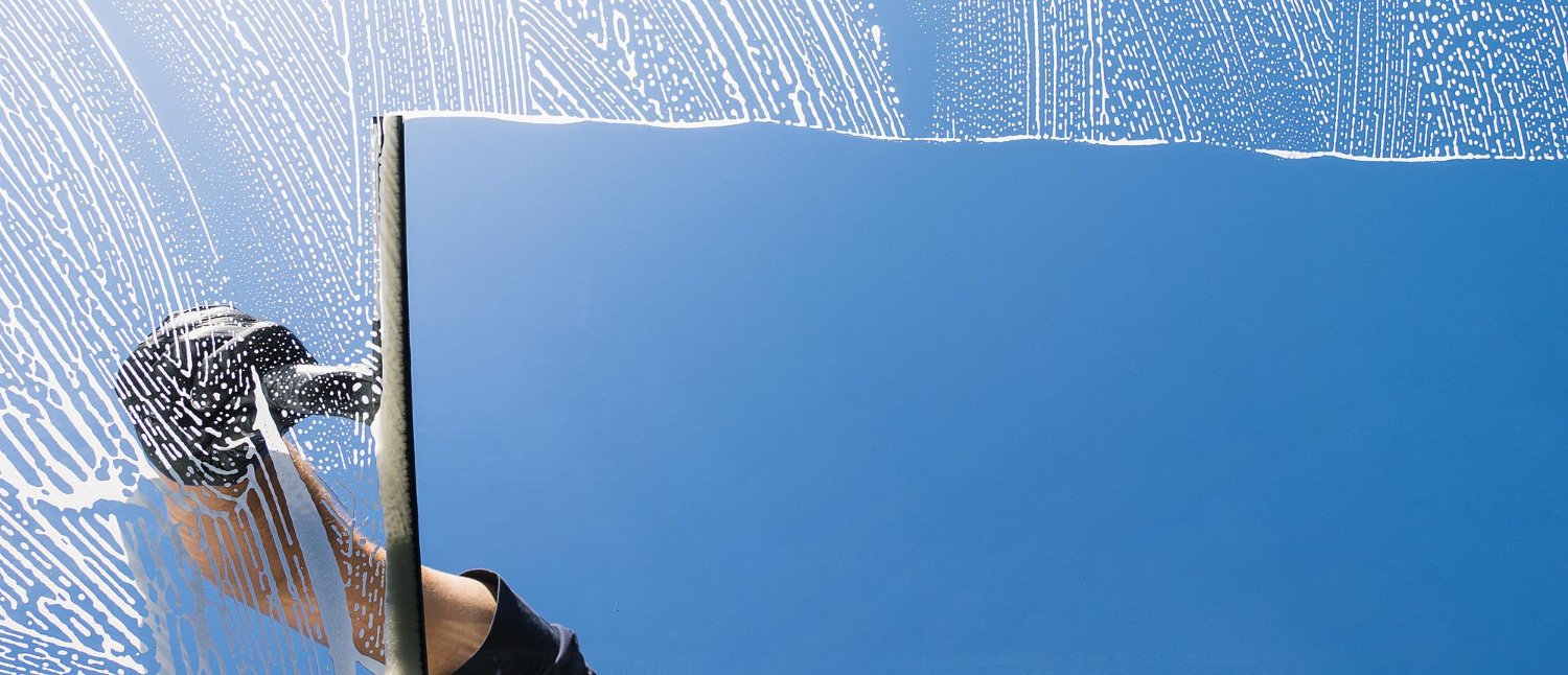 Person wearing gloves wipes a large glass window, with a blue sky in the background.