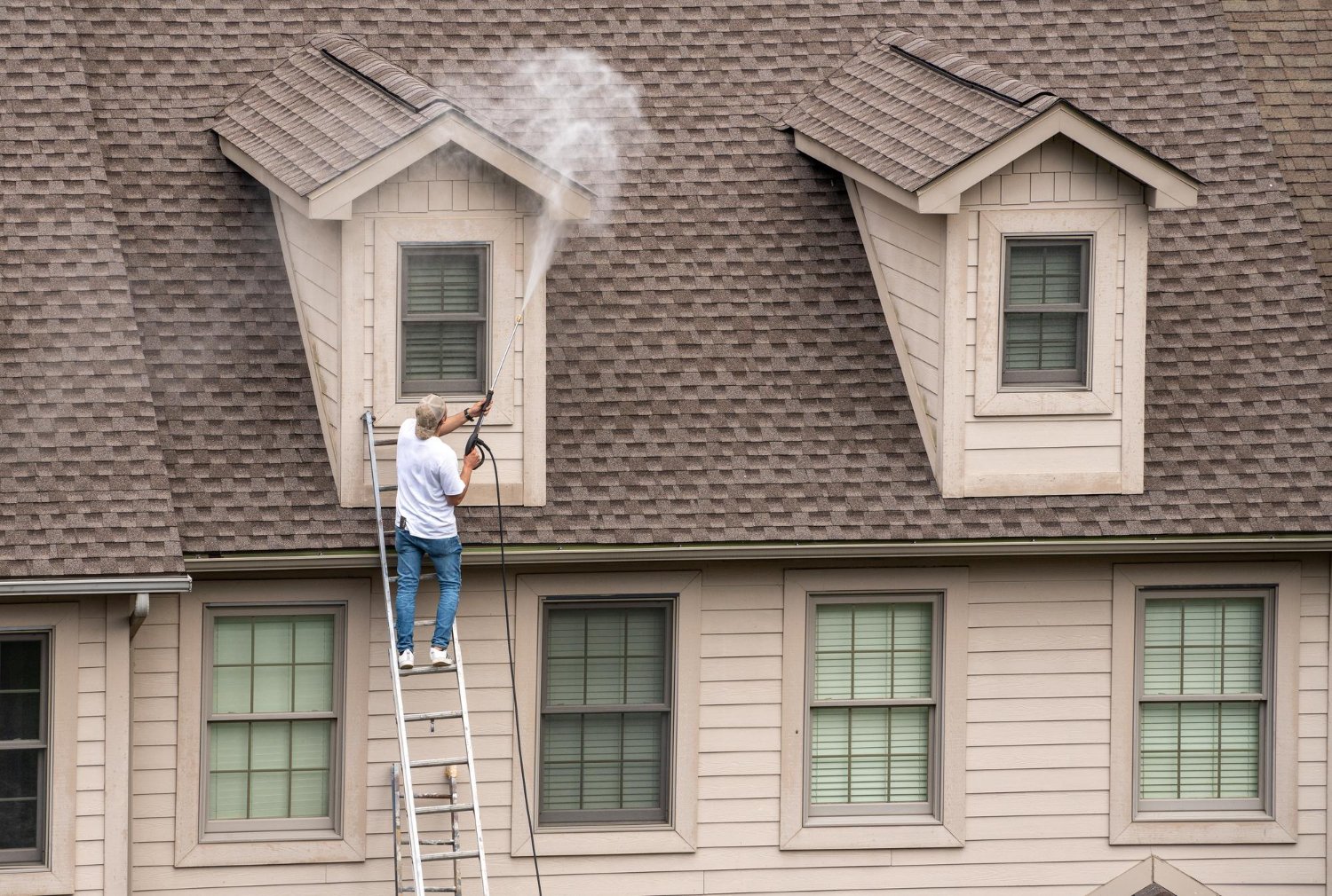 Man on a ladder pressure-washing a dormer window on a brown shingled roof.