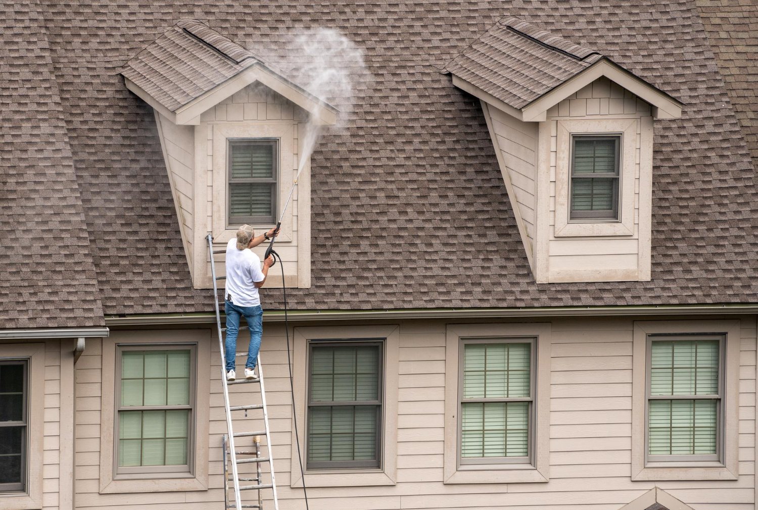 Man on a tall ladder power-washing the roof of a beige two-story house, steam and water spray visible around a dormer window on the left side